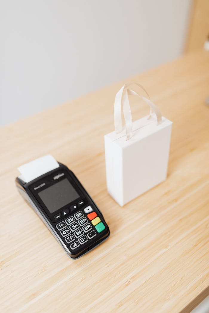 A payment terminal and a small white gift bag on a wooden countertop, ideal for retail or shopping themes.